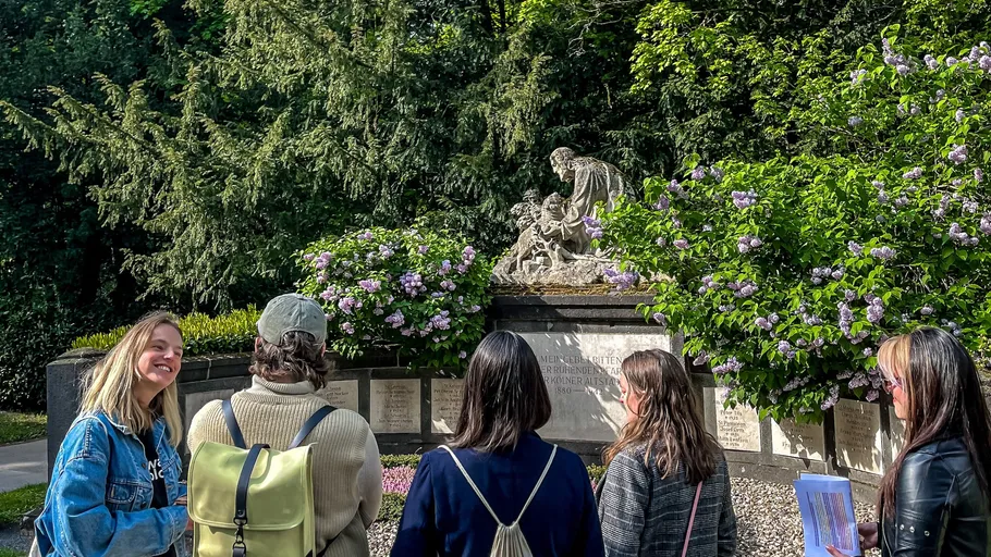 Fünf Personen betrachten eine Friedhofsskulptur im Freien.