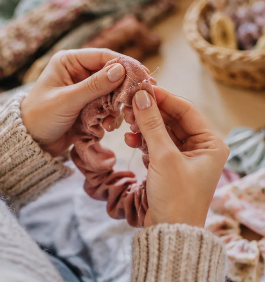 Hands sewing a pink scrunchie indoors.