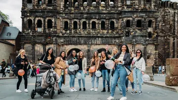 Gruppe Frauen mit Ballons vor antikem Torbogen.