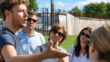 Man speaking to group outdoors near wall.