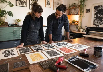Two artists admire prints on a studio table.