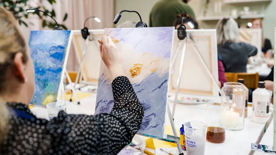 Person painting landscape on canvas in group setting.