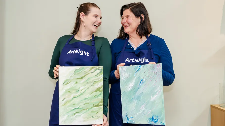 Two women smiling, holding paintings indoors.