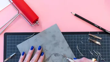 Hands carving linoleum on pink background.