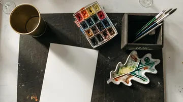 Wasserfarben, Papier und Pinsel auf einem Tisch.