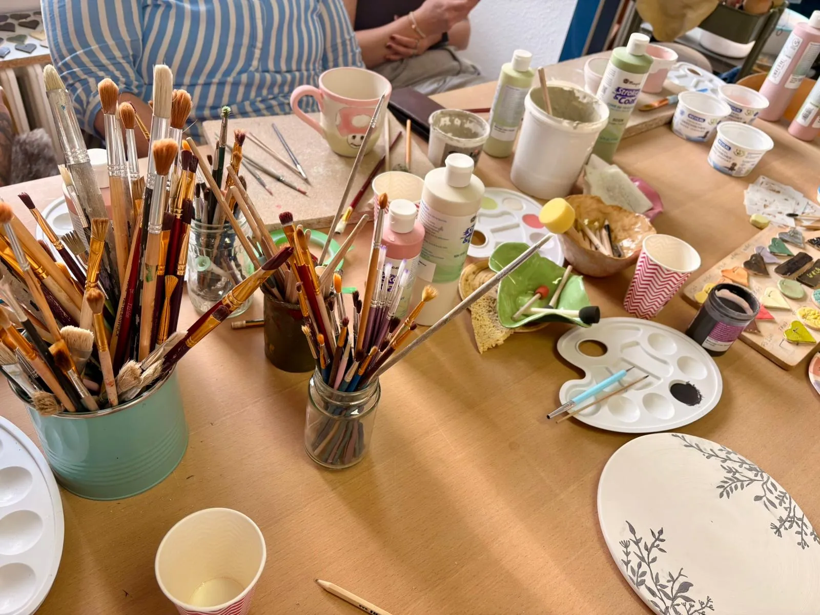 Kunstmaterialien auf Tisch, Personen sitzen daneben.