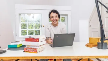 Person lächelt am Schreibtisch mit Büchern und Laptop.