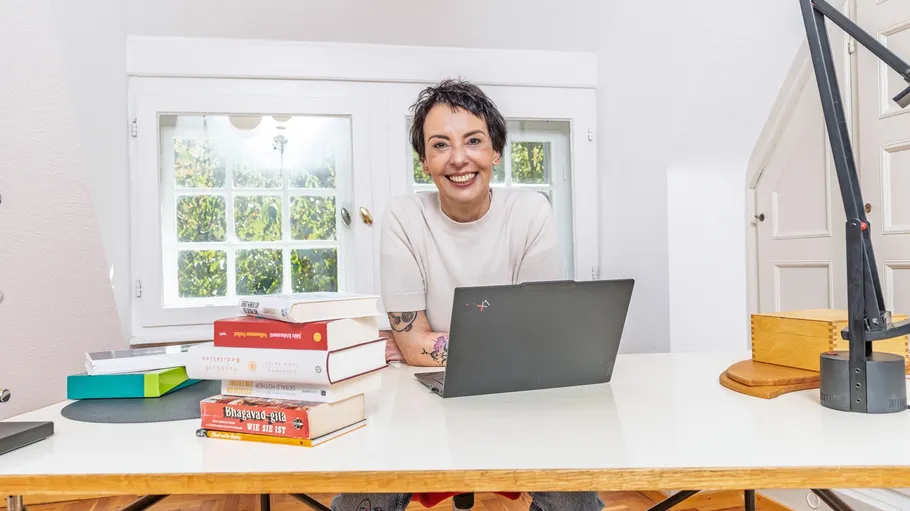 Person lächelt am Schreibtisch mit Büchern und Laptop.