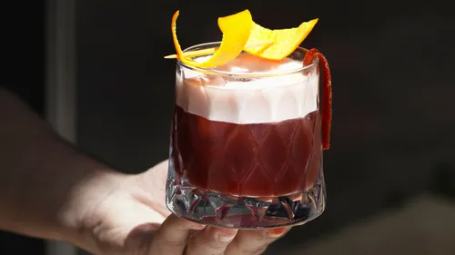 Hand hält Cocktail mit Orangengarnitur.
