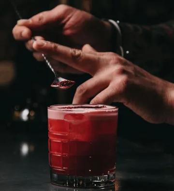 Hand sprinkles red powder on cocktail glass.