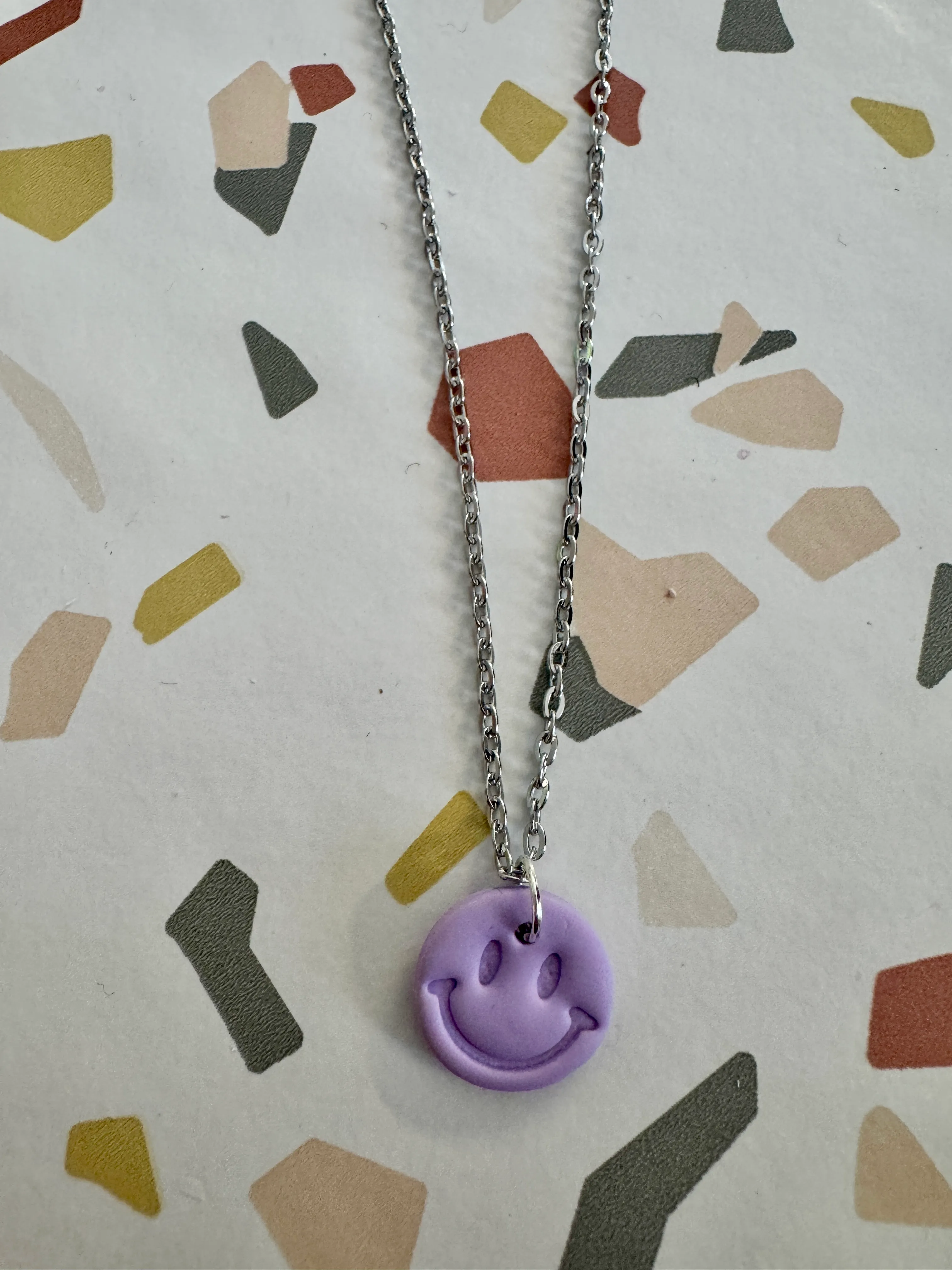 Purple smiley pendant on silver chain, geometric background.