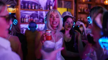Woman with headphones holds drink in busy bar.