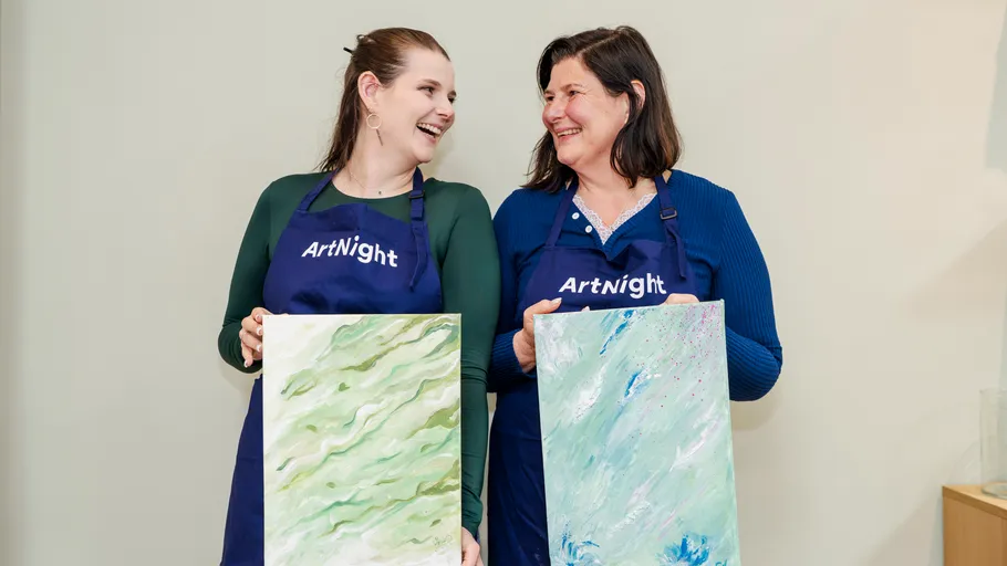 Two women holding paintings, smiling indoors.