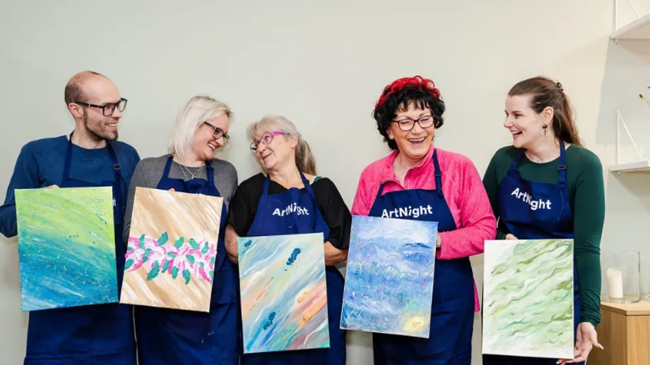 Group holding paintings, wearing aprons, smiling indoors.