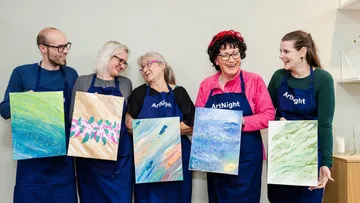 Five people holding paintings, smiling in a studio.