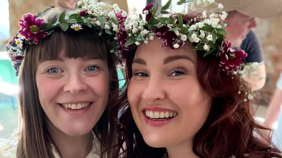 Two women smiling with flower crowns indoors.