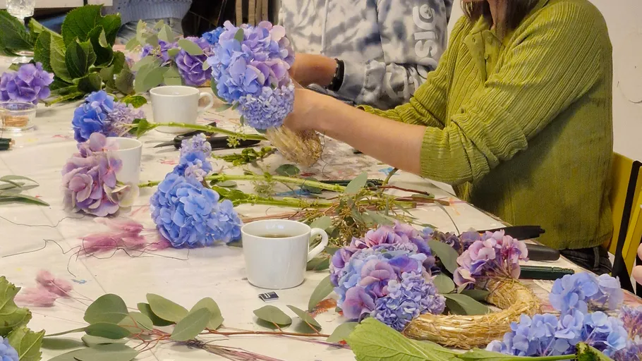 People making wreaths with purple flowers indoors.
