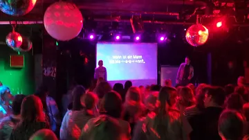 Publikum schaut Karaoke auf Bühne mit Discokugeln.