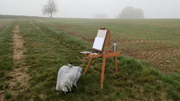 Staffelei und Decke in nebliger Feldlandschaft.
