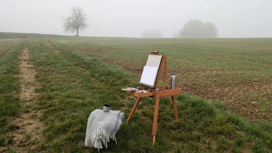 Staffelei und Decke in nebliger Feldlandschaft.