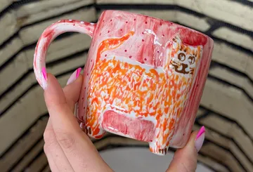 Hand hält katzenbemalten Becher mit pinken Nägeln.