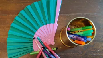 Two handheld fans next to pens on table.