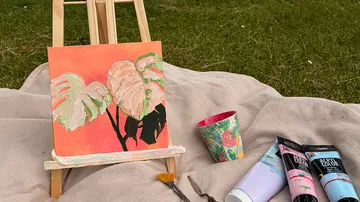 Painting on easel, blanket on grass, art supplies.