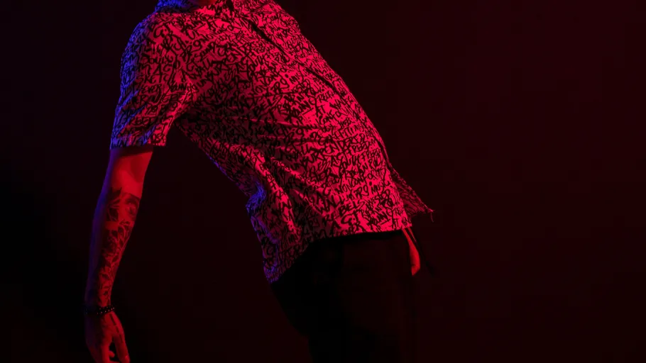 Person leans back wearing patterned shirt, dark room.