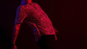 Person leaning in patterned shirt, dark background.