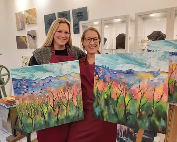 Two women holding landscape paintings indoors.