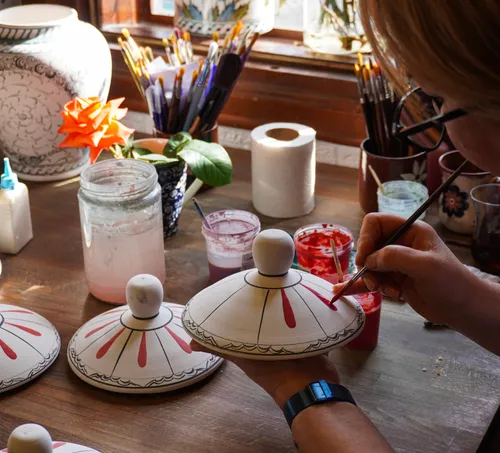 Person painting pottery lid, surrounded by art supplies.