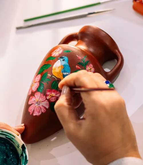 Hands painting colorful bird on vase.
