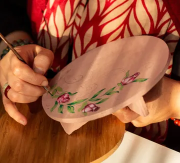Person painting flowers on round wooden object.