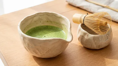 Keramikschüssel mit Matcha und Besen auf Tisch.