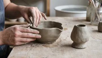 Hände formen Tonschüssel auf Holztisch.