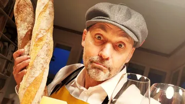 Man holding baguettes indoors, wearing a cap.