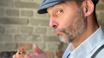 Mann hält Mini-Figur vor Ziegelwand.