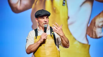 Man speaking on stage wearing a yellow apron.