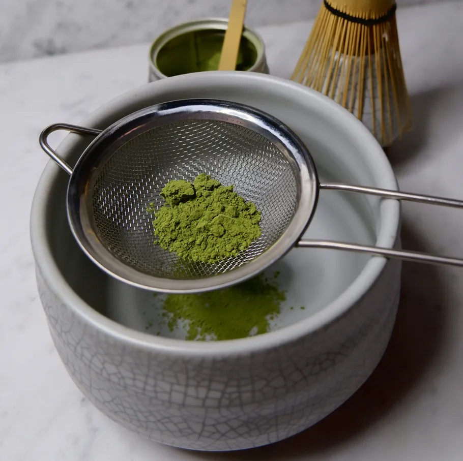 Matcha-Pulver im Sieb über Schale, Besen daneben.