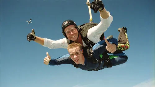 Zwei Personen beim Fallschirmspringen, Daumen hoch, klarer Himmel.