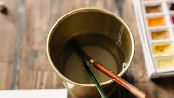 Pinsel im Wasserglas, Aquarellkasten daneben.