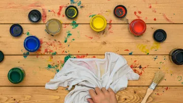 Paint jars open, hand cleaning cloth on wooden table.
