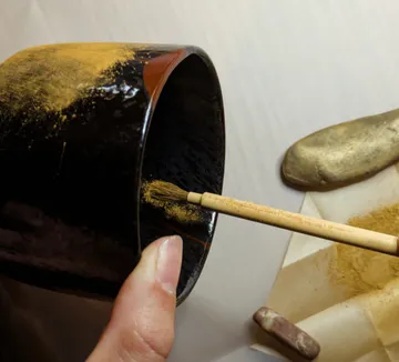 Hand trägt Goldpulver auf schwarze Keramik auf.