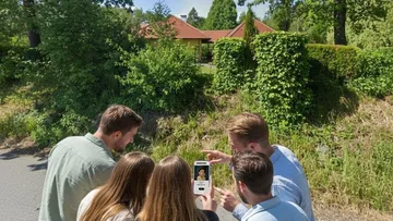 Group examines phone image near house outdoors.