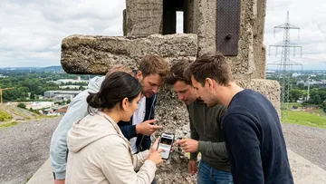 Gruppe untersucht Handy nahe großer Steinkonstruktion.
