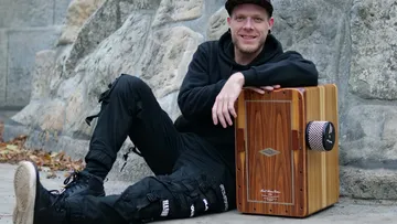 Mann sitzt mit Cajón draußen an Steinwand.