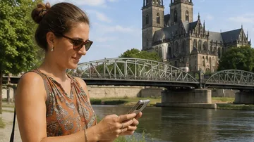 Frau nutzt Handy am Fluss, Dom im Hintergrund.