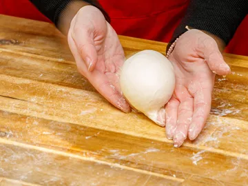 Hände formen Teigball auf einer bemehlten Holzoberfläche.