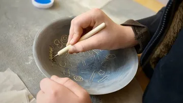 Hands carving patterns on a clay bowl.