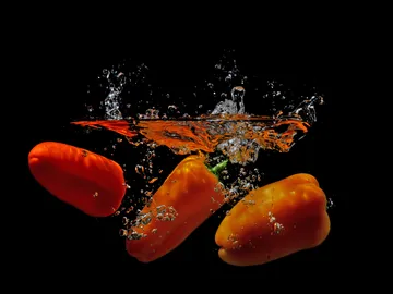 Peppers dip in water, splashing bubbles, black background.